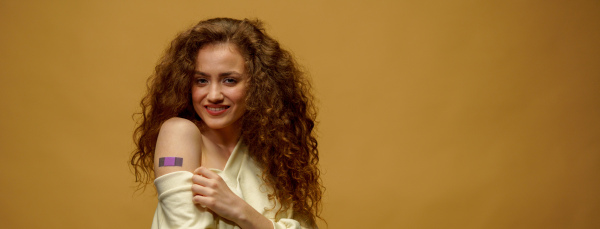 A portrait of a female smiling after getting a vaccine. Woman holding down her shirt sleeve and showing her arm with bandage after receiving vaccination isolated on yellow background.