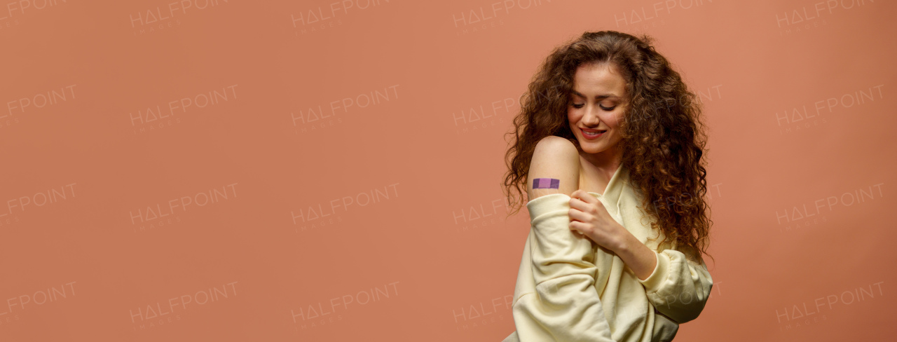 A portrait of a female smiling after getting a vaccine. Woman holding down her shirt sleeve and showing her arm with bandage after receiving vaccination isolated on orange background.