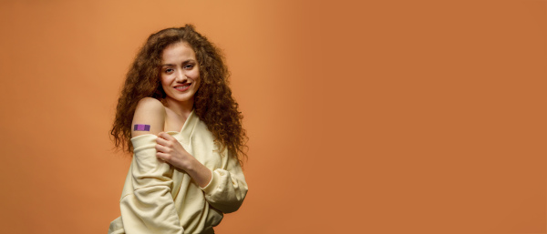 A portrait of a female smiling after getting a vaccine. Woman holding down her shirt sleeve and showing her arm with bandage after receiving vaccination isolated on orange background.