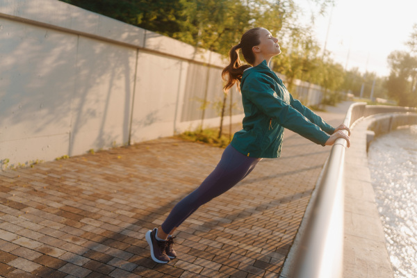 Young female runner stretching arms before her early morning run. Fitness girl in sportswear preparing for evening exercise. Outdoor workout concept.