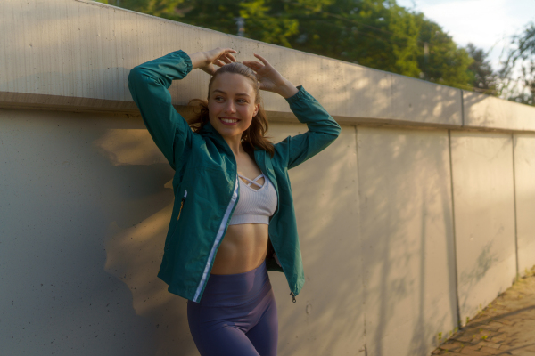 Portrait of young sporty woman in open windbreaker and sports bra. Fitness woman with crossed arms in front of concrete wall in the city. Healthy lifestyle concept.