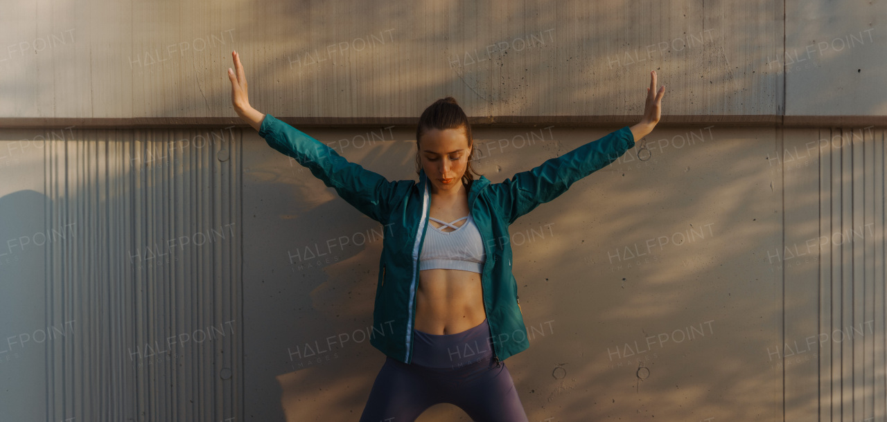 Young female runner stretching and doing deep breathing exercises before her early morning run. Fitness girl in sportswear preparing for evening exercise. Outdoor workout concept.