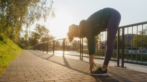 Young female runner stretching arms and legs before her early morning run in the city. Fitness girl in sportswear preparing for evening exercise. Outdoor workout concept. Sillhouette with morning light.