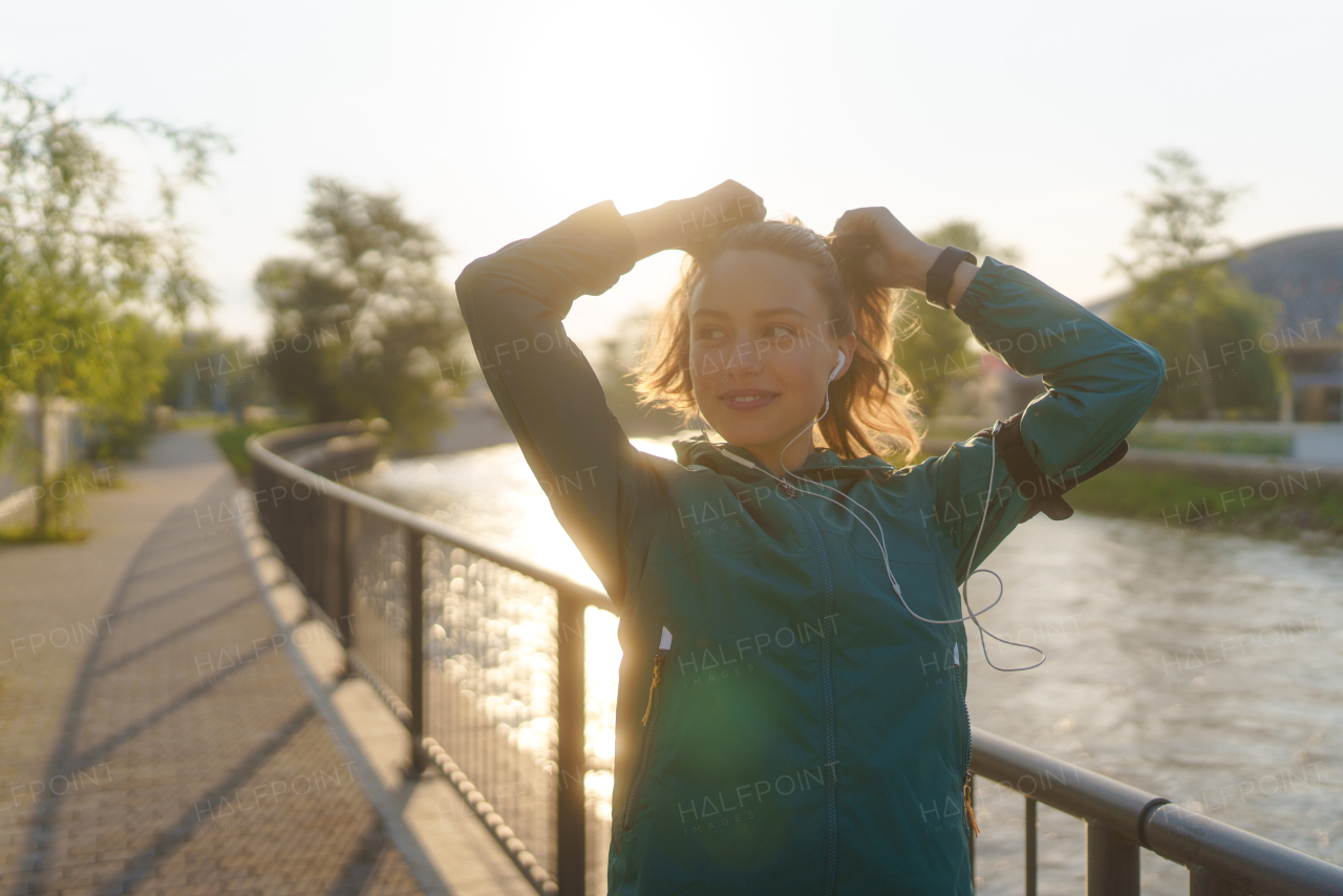 Portrait of young female runner doing workout hairstyle before her early morning run. Fitness girl in sportswear preparing for evening exercise. Outdoor workout concept.