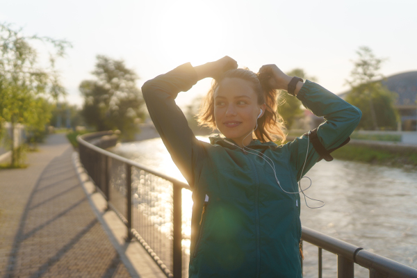 Portrait of young female runner doing workout hairstyle before her early morning run. Fitness girl in sportswear preparing for evening exercise. Outdoor workout concept.