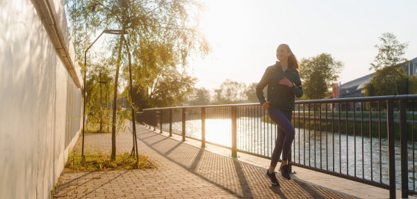 Athletic young woman in activewear running along the embankment of river at the morning. Outdoor workout concept.