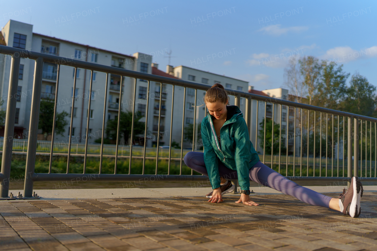 Young female runner stretching before her early morning run in the city. Fitness girl in sportswear preparing for evening exercise. Outdoor workout concept.