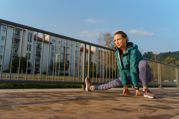 Young female runner stretching legs before her early morning run in the city. Fitness girl in sportswear preparing for evening exercise. Outdoor workout concept.