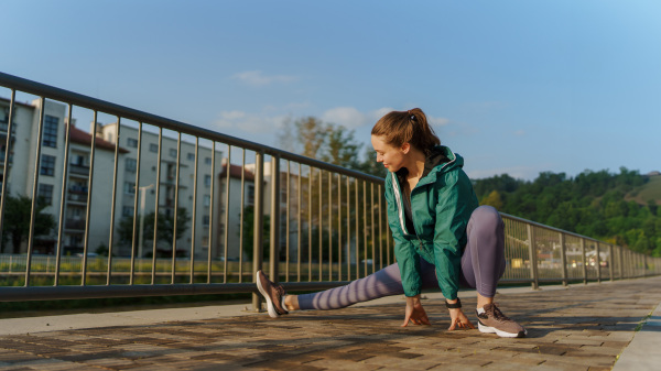 Young female runner stretching legs before her early morning run in the city. Fitness girl in sportswear preparing for evening exercise. Outdoor workout concept.