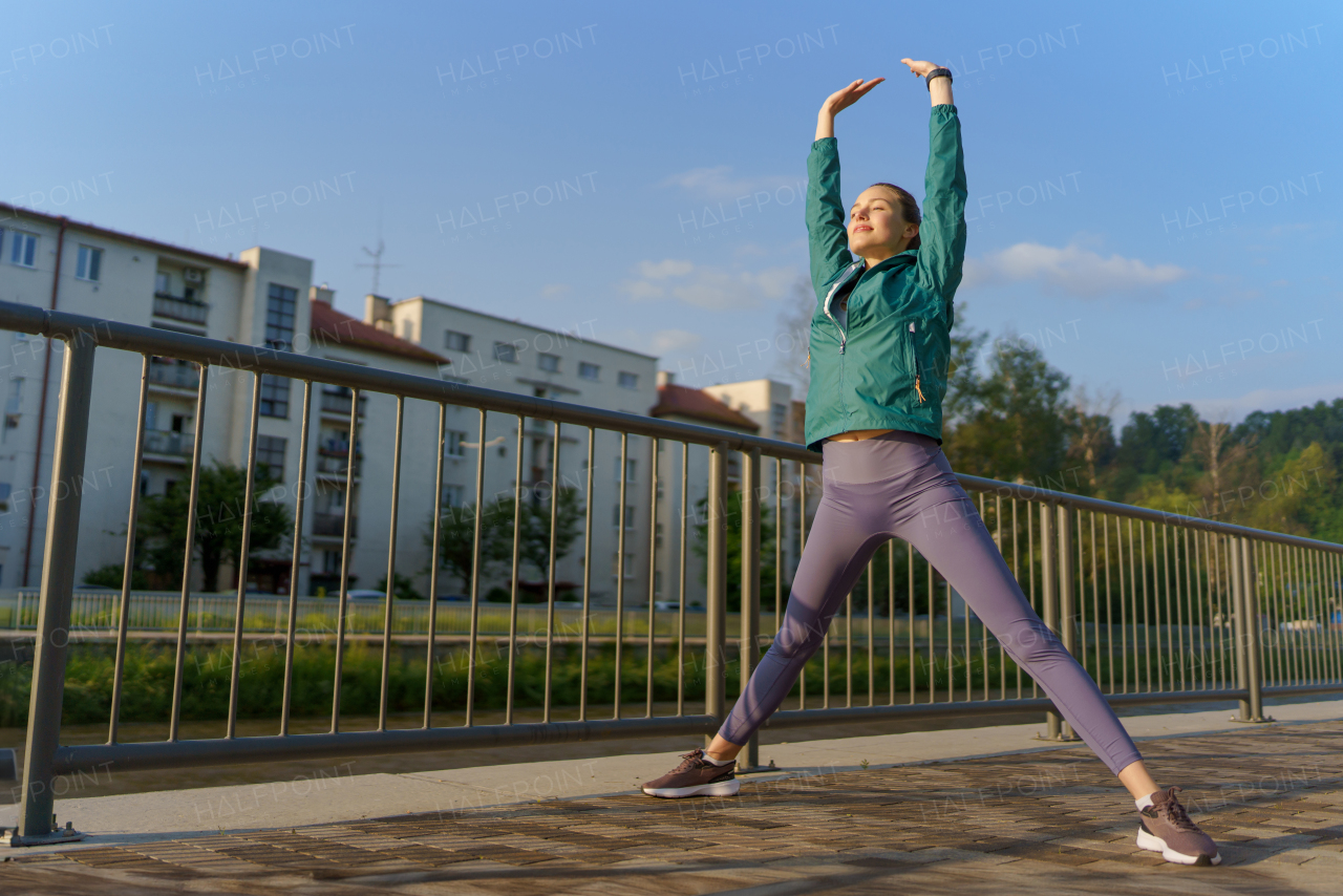 Young female runner stretching before her early morning run in the city. Fitness girl in sportswear preparing for evening exercise. Outdoor workout concept.