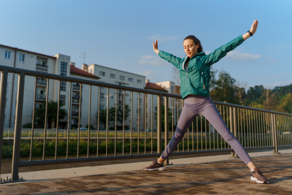 Young female runner stretching arms and legs before her early morning run in the city. Fitness girl in sportswear preparing for evening exercise. Outdoor workout concept.