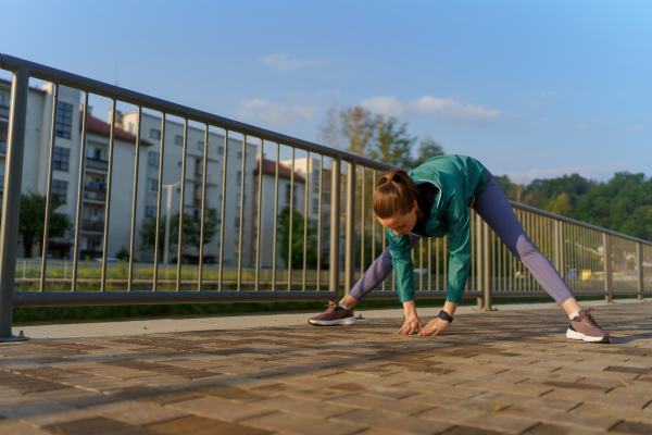 Young female runner stretching before her early morning run in the city. Fitness girl in sportswear preparing for evening exercise. Outdoor workout concept.