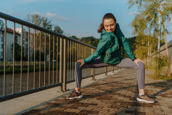 Young female runner stretching before her early morning run. Fitness girl in sportswear preparing for evening exercise. Outdoor workout concept.