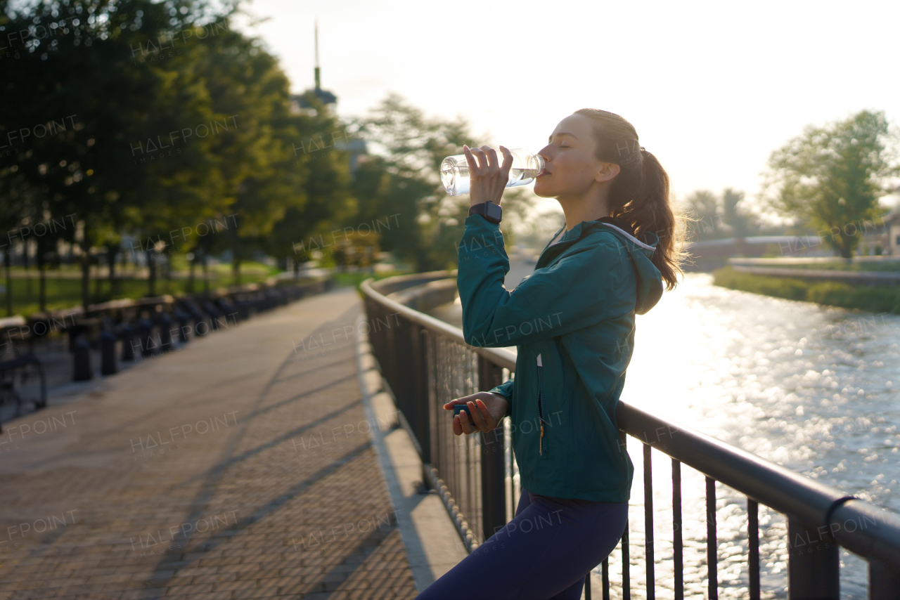 Young female runner drinking water from bottle before her early morning run in the city. Fitness girl in sportswear rehydrating after evening exercise. Outdoor workout concept.