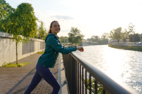 Side view of young female runner stretching before her early morning run. Fitness girl in sportswear preparing for evening exercise. Deep breathing exercises before workout.