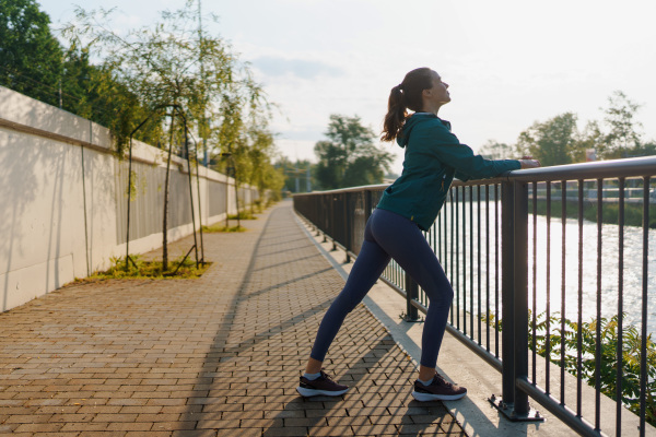 Side view of young female runner stretching before her early morning run. Fitness girl in sportswear preparing for evening exercise. Deep breathing exercises before workout.