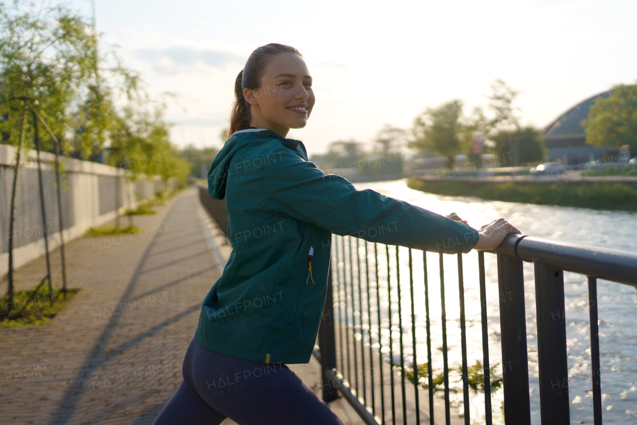 Young female runner stretching before her early morning run in the city. Fitness girl in sportswear preparing for evening exercise. Outdoor workout concept.