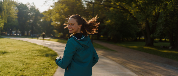 Portrait of young beautiful female runner in blue jacket with wind blowing up her hair jogging across the city park. Evening workout in the city. Outdoor workout concept.