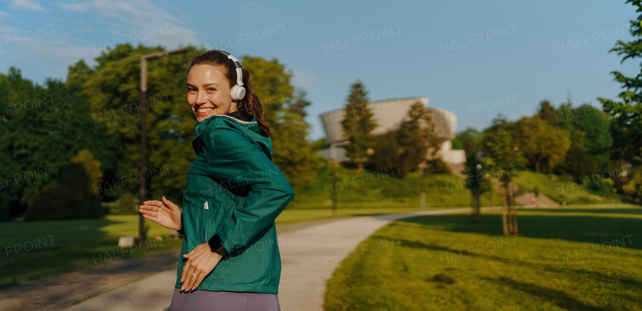 Portrait of young beautiful female runner with headphones with wind blowing up her hair jogging across the city park. Evening workout in the city. Outdoor workout concept.