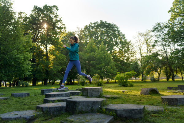 Full lenght portrait of beautiful fitness woman jumping on concrete obstacles in the park. Training for obstacle course, adventure racing. Workout at the park.