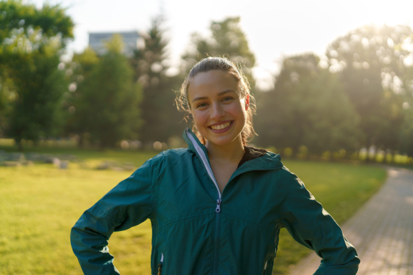 Portrait of young beautiful female runner in blue jacket in the city park. Evening workout in the city. Outdoor workout concept.