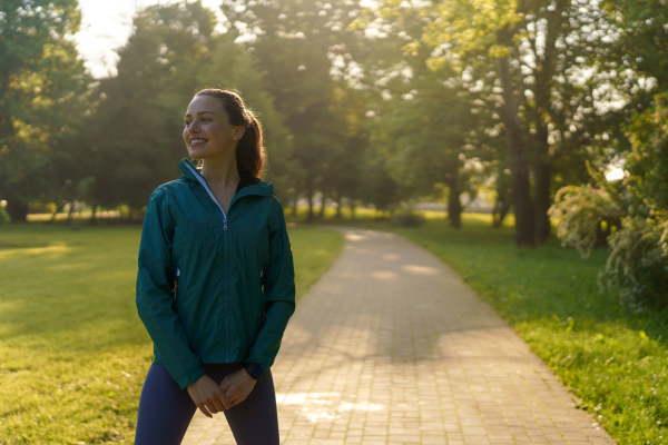 Portrait of young beautiful female runner in blue jacket with wind blowing up her hair jogging across the city park. Evening workout in the city. Outdoor workout concept.