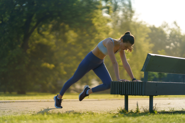 Full lenght portrait of beautiful fitness woman doing mountain clibers exercise against park bench. Workout at the park. Elevated mountain climbers.