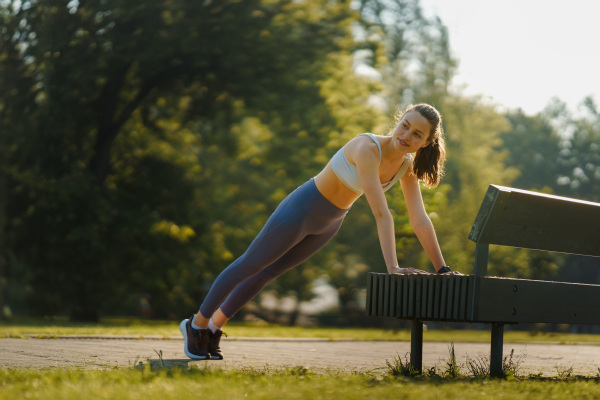 Full lenght portrait of beautiful fitness woman doing push-ups against park bench. Workout at the park. Park bench excercises.