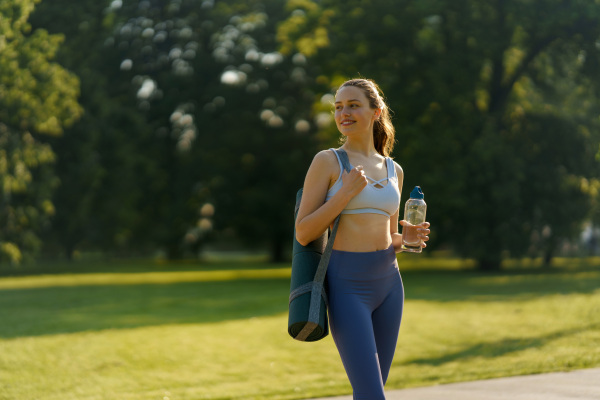 Medium shot of athletic gir in sportswear with yoga mat on shoulder and water bottle heading for a pilates workout.