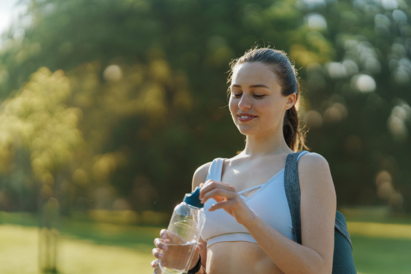 Medium shot of athletic gir in sportswear with yoga mat on shoulder drinking from reusable water bottle.