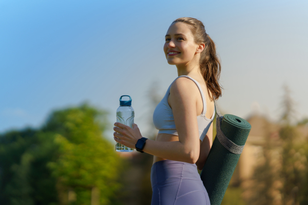 Side view of beautiful athletic girl in sportswear with yoga mat on shoulder and water bottle heading for a pilates workout.