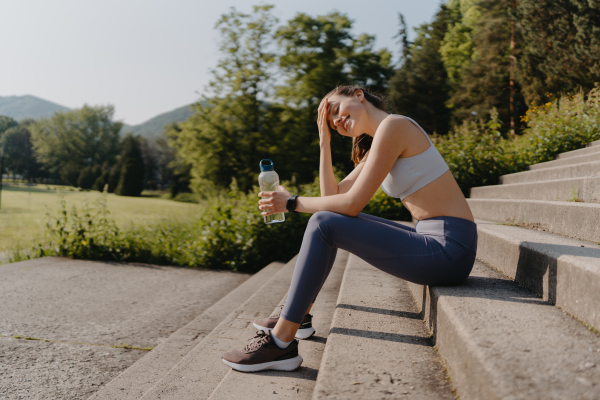 Side view of young fitness woman in sportswear resting after workout session in the city. Beautiful sporty woman drinking water on concrete stairs after morning excercise.