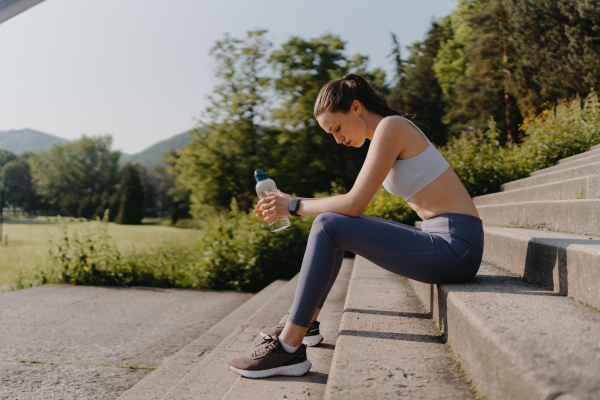 Side view of exhausted fitness woman in sportswear resting after workout session in the city. Beautiful sporty woman drinking water on concrete stairs after morning excercise.