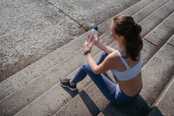 Top view of exhausted fitness woman in sportswear resting after workout session in the city. Beautiful sporty woman drinking water on concrete stairs after morning excercise.