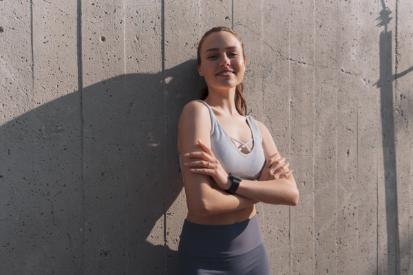 Portrait of young sporty woman in sportswear standing with crossed arms outdoors. Fitness woman in front of concrete wall in the city. Healthy lifestyle concept. Street workout.