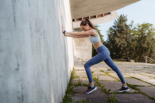 Side view of young sporty woman in sportswear excercising outdoors. Fitness woman stretching against concrete wall in the city. Healthy lifestyle concept. Street workout.