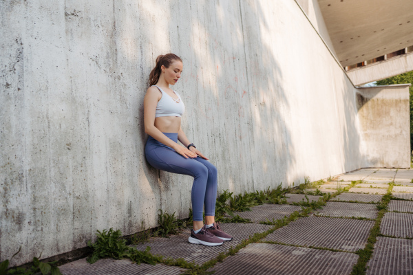 Portrait of young sporty woman in sportswear excercising outdoors, doing wall sit exercise. Fitness woman in front of concrete wall in the city. Street workout.