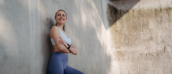 Portrait of young sporty woman in sportswear resting after excercising outdoors. Fitness woman in front of concrete wall in the city. Banner with copy space. Healthy lifestyle concept.