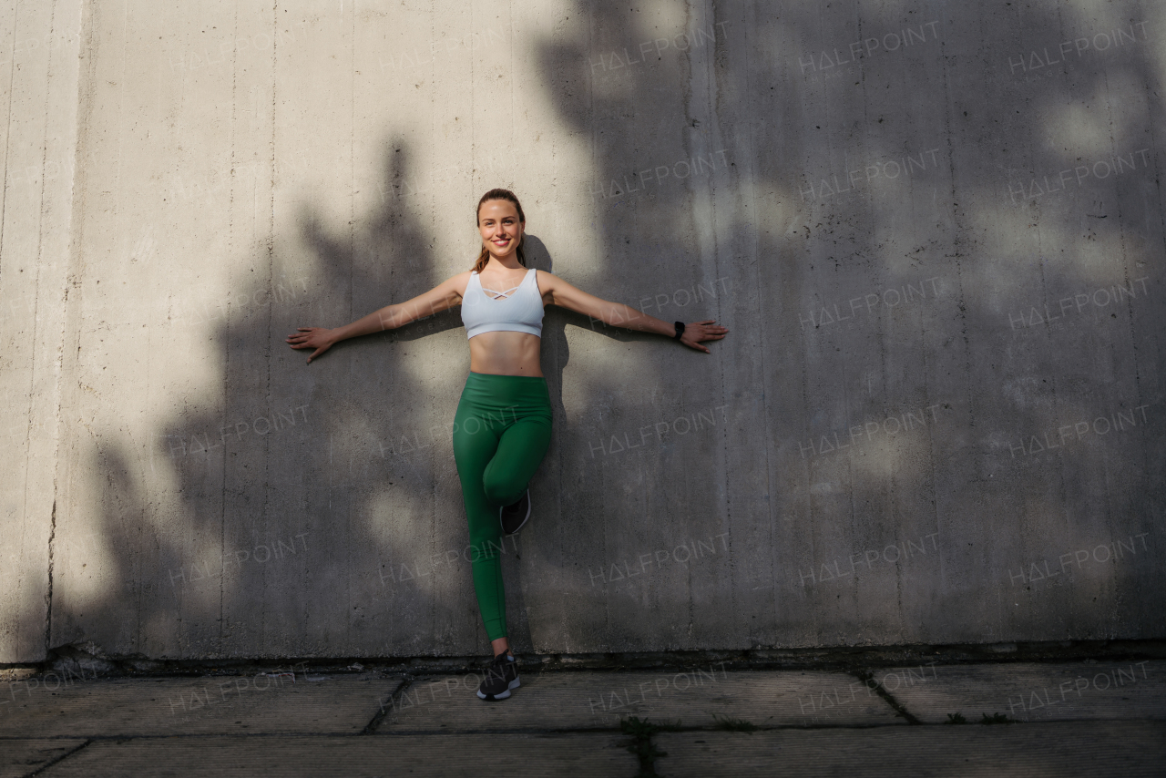 Portrait of young sporty woman in sportswear leaning against the wall. Smiling woman in front of concrete wall in the city. Healthy lifestyle concept. Street workout.