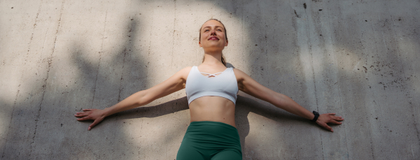 Portrait of young sporty woman in sportswear standing with opened arms outdoors. Fitness woman in front of concrete wall in the city. Healthy lifestyle concept. Banner with copyspace on gray bacground.