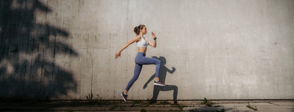 Beatutiful female athlete in sportswear exercising in the city. Running in front of concrete wall casting shadow. Banner with copy space.