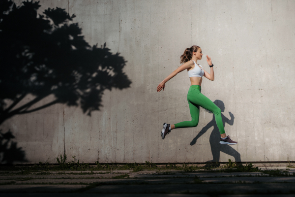 Female athlete in sportswear exercising in the city. Running in front of concrete wall casting shadow. Banner with copy space.