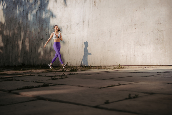 Female athlete in sportswear exercising in the city. Running in front of concrete wall casting shadow. Banner with copy space.