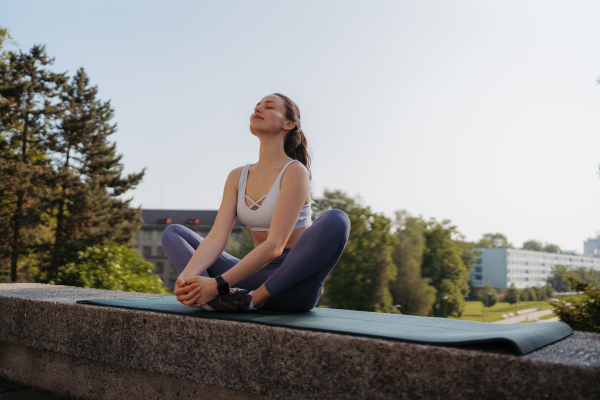 Front view of young sporty woman doing breathing exercises on gym mat after workout in the city. Woman meditate outdoors, in the park.