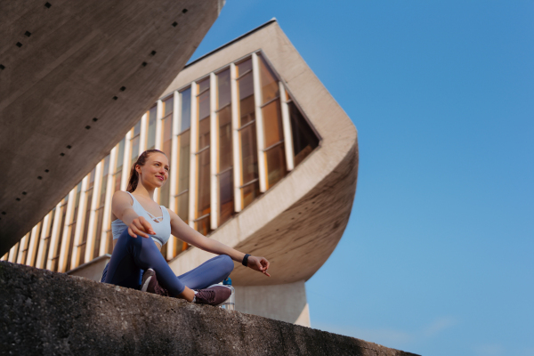 Low angle shot of young sporty woman doing breathing exercises after workout in the city. Woman meditate outdoors, sitting on concrete stairs in the park.