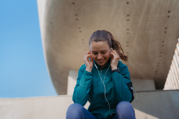 Young fitness woman listening to music with headphones, resting after workout session in the city. Beautiful sporty woman enjoying sunrise after morning excercise.