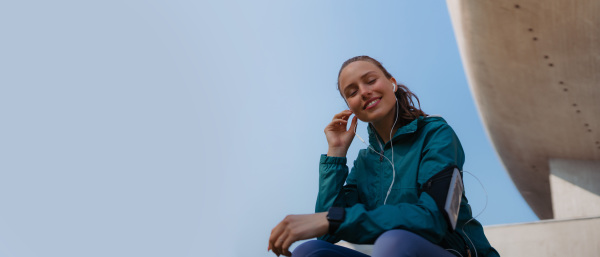 Young fitness woman listening to music with headphones, resting after workout session in the city. Beautiful sporty woman enjoying sunrise after morning excercise. Banner with copyspace.