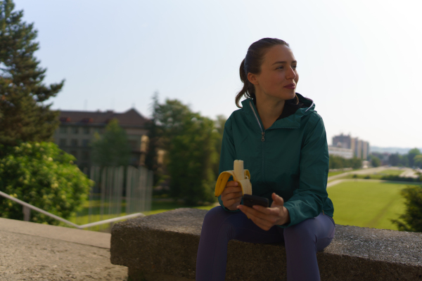 Portrait of athletic girl in sportswear eating banana and checking her smartphone after workout.