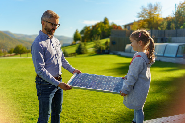 Father with his little daughter catching sun at solar panel,charging at the backyard. Alternative energy, saving resources and sustainable lifestyle concept.