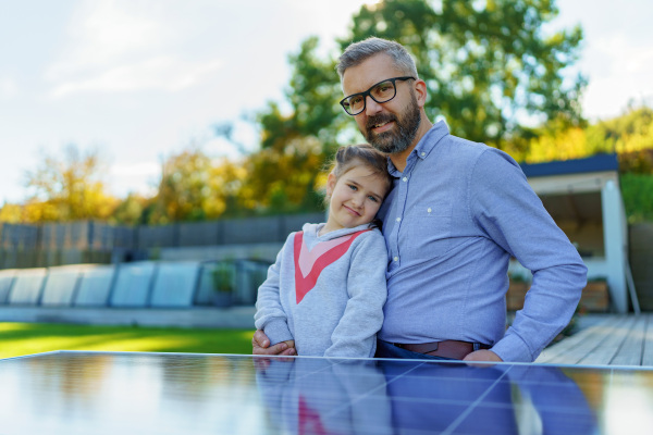 Father with his little daughter catching sun at solar panel,charging at the backyard. Alternative energy, saving resources and sustainable lifestyle concept.